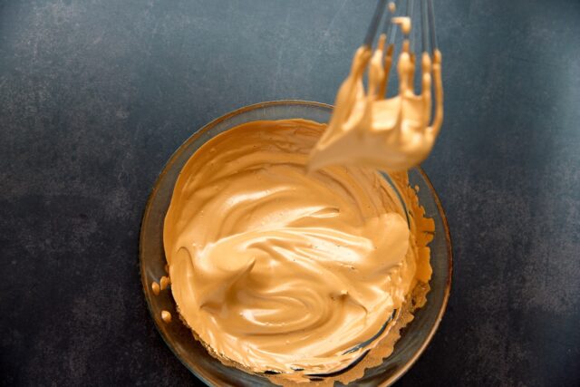 Close up of glass bowl and whisk with fluffy creamy whipped coffee foam on a dark background. Trendy food and drink concept. Copy space.